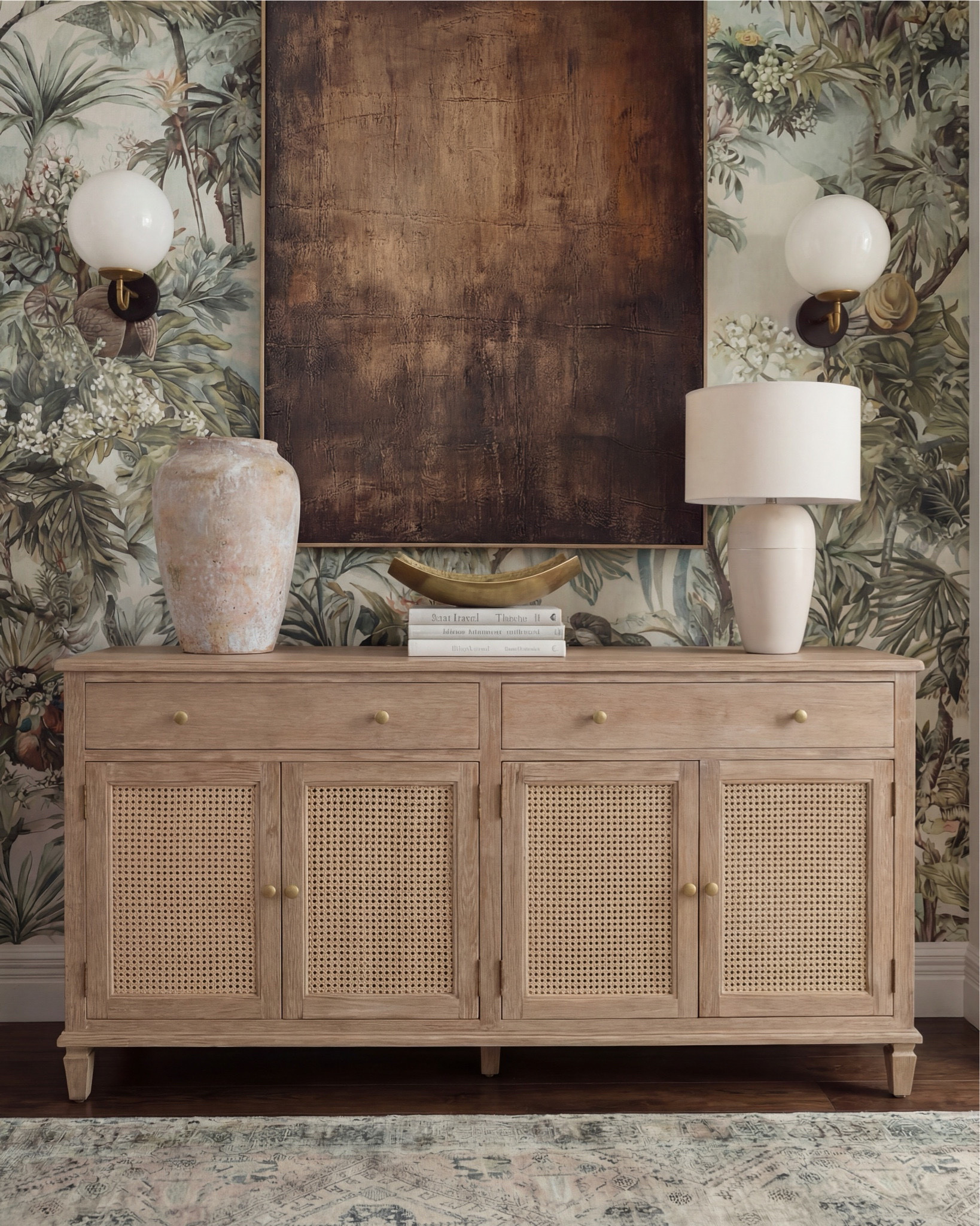 There’s something about a moody, nature-inspired foyer that just sets the tone for the whole home, and this one does it so effortlessly. The mix of warm wood, soft texture, and bold art makes the space feel collected and elevated from the moment you walk in.

The natural-oak console brings the perfect balance of clean lines and subtle detail with its woven cane doors. I styled it with an oversized stone vase, a tall ceramic lamp, and a sculptural gold bowl layered on books — just enough to feel intentional without being cluttered.

The vintage-inspired runner adds muted pattern and softness underfoot, grounding all the warm wood tones beautifully. And that wallpaper — the deep greens, palms, and floral textures make the entire wall feel like a piece of art on its own.

The oversized copper-toned artwork brings so much richness to the scene, pulling in the darker notes from the sconces and the natural shadows in the wallpaper. It’s the kind of statement piece that instantly elevates a foyer.

✨ Shop the Look
Natural-oak cane console | Oversized stone vase | Sculptural gold bowl | Ceramic table lamp | Botanical wallpaper | Vintage-style runner | Copper-toned oversized artwork | Globe wall sconces

#LTKsale #LTKhome #LTKstyletip