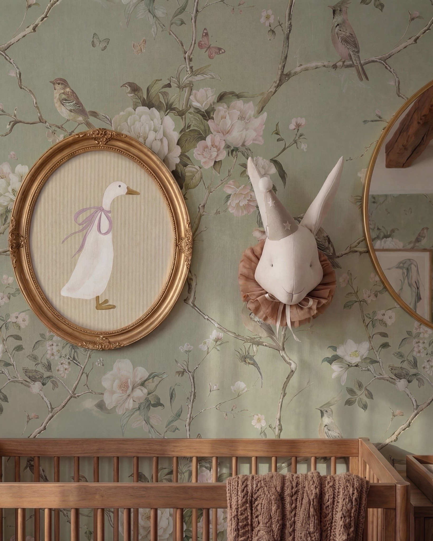 This nursery feels storybook-soft and timeless, blending nature-inspired charm with warm, grounding materials. The floral wallpaper sets a whimsical tone, while exposed wood beams and rich wood furniture keep the space feeling elevated and cozy rather than overly sweet.

The balance of classic nursery pieces with refined finishes—like brass accents, woven textures, and sculptural lighting—makes this room feel calm, intentional, and designed to grow with the child.

Shop the Look:
• Floral wallpaper (soft green with botanical print)
• Solid wood crib (warm walnut tone)
• Knit throw blanket (neutral brown)
• Wood dresser with woven drawer fronts
• Changing tray (wood)
• Round brass-framed wall mirror
• Oval framed animal artwork
• Decorative wall animal mount
• Upholstered accent chair (ivory)
• Green accent pillows
• Floor lamp (brass base, white shade)
• Table lamp (ceramic base, white shade)
• Pendant ceiling light (soft white globe)
• Neutral vintage-style area rug
• Light wood flooring
• Sheer curtains with linen drapery panels
#LTKSaleAlert

#LTKsale #LTKhome