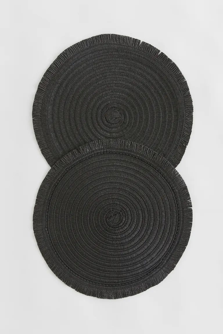 A pair of round place mats in braided paper straw with a fringed trim all around. | H&M (US)