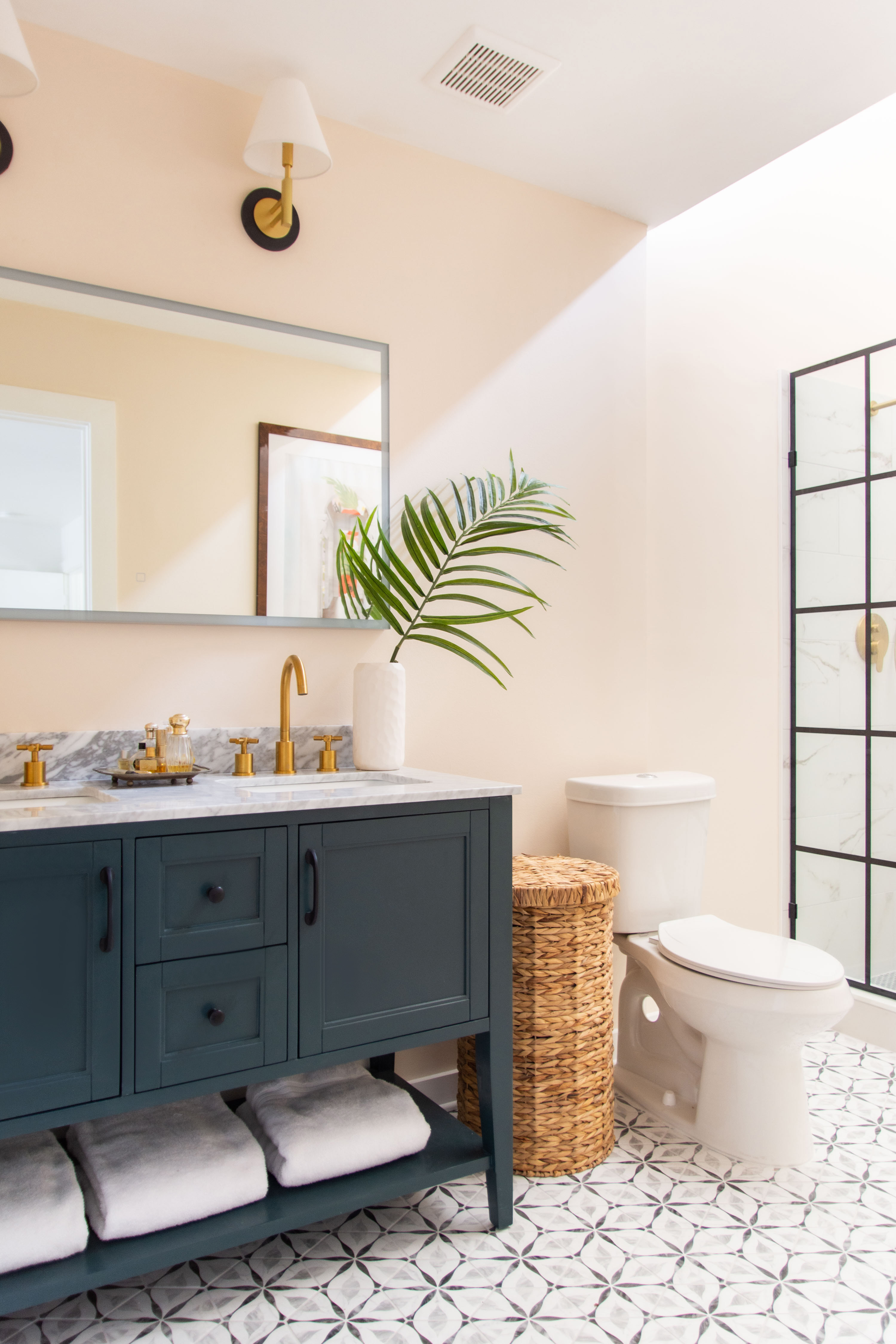 Modern bathroom design goals. This space combines a navy blue vanity with gold hardware, marble countertops, and a sleek double sink. The patterned tile floor adds personality, while the wicker laundry hamper and palm leaf decor bring in warmth and texture. A walk-in shower with black framed glass doors gives a clean, contemporary touch. Perfect inspiration for anyone looking for primary bathroom ideas, vanity styling tips, or timeless bathroom remodel inspiration. 

 