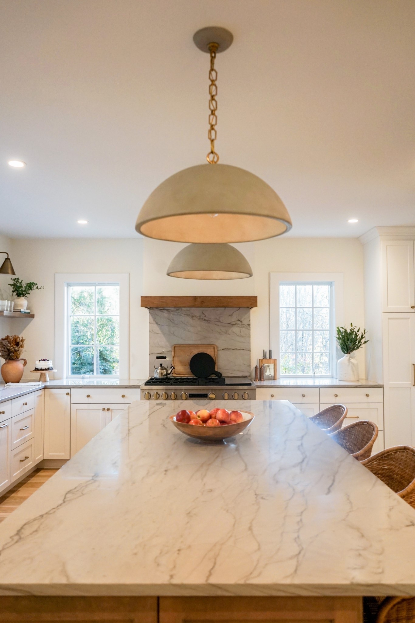 A beautifully designed kitchen featuring a large marble island with elegant veining, warm rattan barstools, and modern dome pendants... The space is brightened by natural light while white cabinetry and subtle wood accents create a cozy, sophisticated atmosphere!