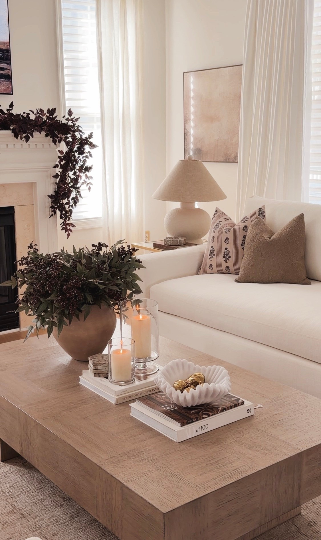 Cozy neutral living room for fall with a white sofa, brown patterned pillows, wood coffee table, glowing candles, autumn greenery and dark fall foliage draped over a white fireplace mantel.