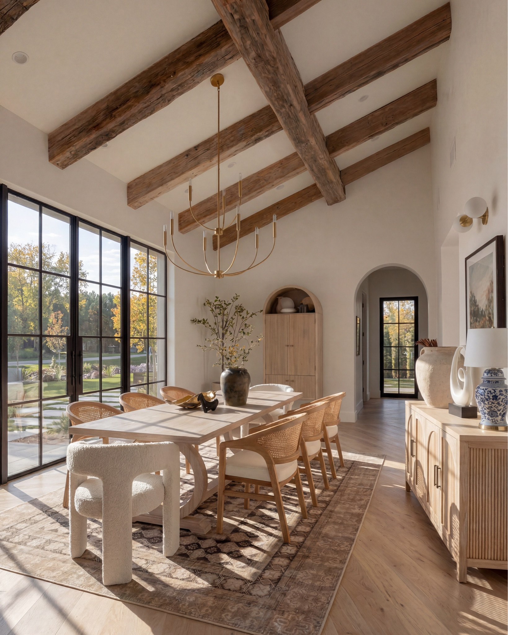 * Rectangular light wood dining table (traditional trestle base)
* Woven cane dining chairs (natural oak frame)
* Neutral vintage-style area rug (distressed pattern)
* Brass modern chandelier (linear / sculptural arms)
* Rustic exposed wood ceiling beams (reclaimed look)
* Large black-framed sliding glass doors (industrial style)
* Dark ceramic vase (handmade / organic texture)
* Dried floral arrangement (neutral stems)
* Sculptural brass decorative bowl
* Black ceramic candle holder (modern abstract form)
* White taper candle
* Small matte black decorative bowl
* Minimalist sculptural object (black accent piece)
* Light wood sideboard cabinet (fluted / ribbed detailing)
* Built-in arched niche cabinet (custom millwork look)
* Decorative pottery (neutral ceramic vases)
* Framed landscape wall art (classic / muted tones)
* Indoor olive tree (or faux olive tree)
* Neutral linen / textured textiles
* Warm light oak flooring
* Soft cream / off-white wall paint
* Arched doorway architectural detail


#LTKHome #LTKSaleAlert