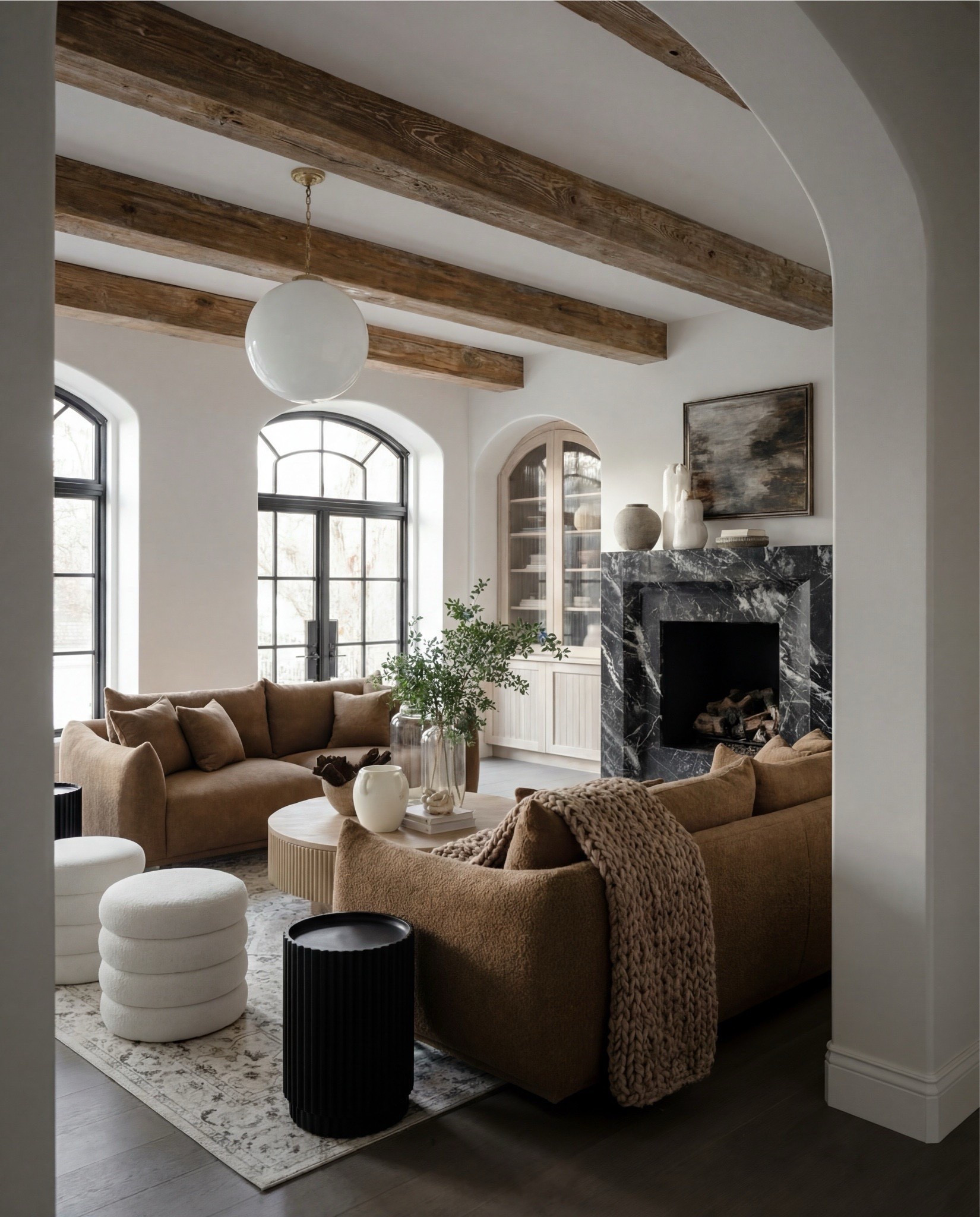 This living room feels grounded, cozy, and effortlessly refined—anchored by rich brown upholstery and dramatic architectural details. The exposed wood ceiling beams add rustic warmth, while the arched doorways and black-framed windows give the space a soft European character. A dark marble fireplace creates a bold focal point, balanced by creamy walls and layered neutral textures.

The styling leans tactile and inviting: plush seating wrapped in textured fabrics, a fluted wood coffee table at the center, and a mix of ceramics and greenery that keeps the room feeling lived-in rather than styled. Knitted throws, soft poufs, and subtle contrast pieces add depth without overpowering the calm palette.

Shop the Look:
• Brown upholstered sofa (textured fabric)
• Brown upholstered accent chair
• Round fluted wood coffee table
• Black ribbed side table
• Neutral vintage-style area rug (cream + taupe)
• White boucle pouf ottomans
• Chunky knit throw blanket (warm neutral)
• White globe pendant light
• Exposed wood ceiling beams
• Arched doorway detail
• Black-framed arched windows
• Dark marble fireplace surround
• Wood built-in cabinet with glass doors
• Ceramic vases (white + stone tones)
• Decorative pottery vessels
• Coffee table books (neutral covers)
• Indoor greenery in glass vase
• Framed abstract artwork (earth tones)


#LTKSaleAlert #LTKHome