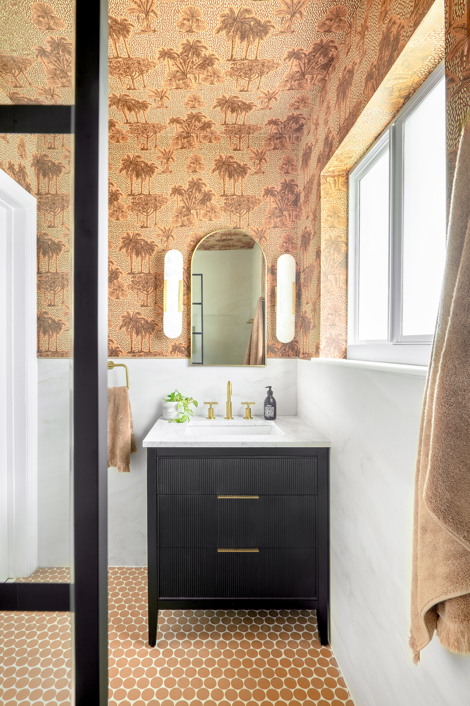 Small space, big personality. This powder bath layers warm tropical wallpaper with a sleek black vanity, brass fixtures, and soft marble surfaces to create a space that feels both bold and timeless. The textured wallpaper brings movement and warmth, while the modern lighting and sculptural mirror keep the look elevated and refined. Save this look for powder room inspiration and shop similar pieces to recreate this statement bathroom design at home. 

 #LTKHome