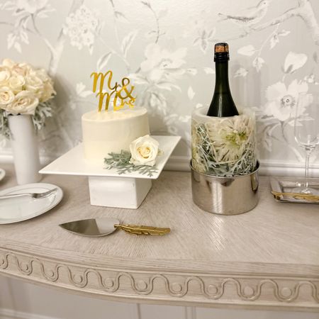 A little diy wedding cake dessert & champagne set up at home on our dining room console table. DIY white rose floral bouquet held in a white vase, cake stand with 6” buttercream cake, and a floral filled ice mold champagne bucket with a gold flower cake knife server & coordinating gold feather tray for the glassware. Traditional, chinoiserie, silver, gold, grandmillenial, French, home decor, entertaining at home  

Follow my shop @HousewifeHospitality on the @shop.LTK app to shop this post and get my exclusive app-only content!

#liketkit #LTKunder100 #LTKwedding #LTKhome
@shop.ltk
https://liketk.it/4eCGT

#LTKhome #LTKwedding #LTKsalealert