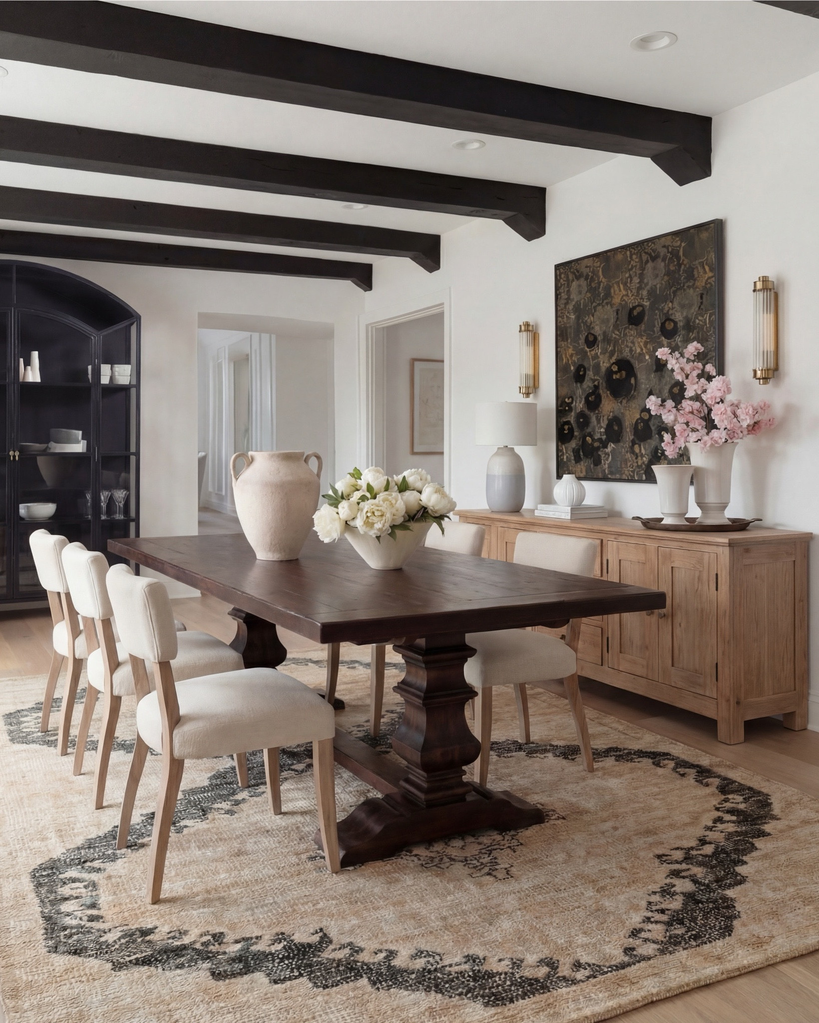Shop the Look:
• Dark wood pedestal dining table
• Cream upholstered dining chairs with light wood legs
• Large neutral area rug with charcoal medallion pattern
• Black arched display cabinet / hutch
• Light wood sideboard / buffet cabinet
• Large ceramic floor vase (neutral tone)
• White ceramic bowl with white florals (peonies or roses)
• Pink faux cherry blossom stems in white vase
• Neutral ceramic table lamp with white shade
• Tall brass wall sconces with glass detailing
• Framed botanical artwork (set of four)
• Dark abstract wall art (large statement piece)
• White decorative vases (assorted sizes)
• Light linen curtains

#LTKhome #LTKsale #LTKstyletip