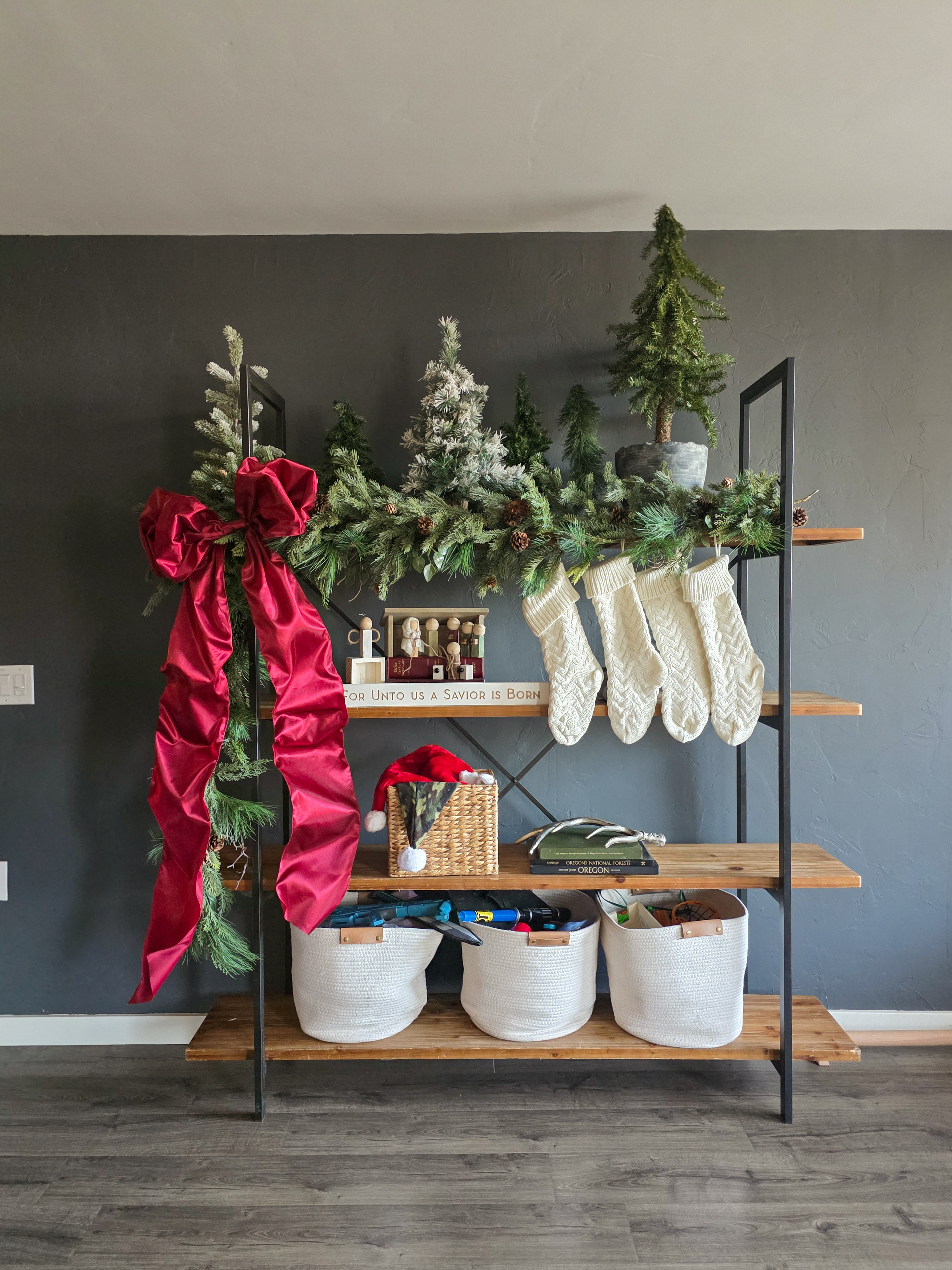 Shelf styling for the holidays: Layered faux trees, knit stockings, and a wooden nativity scene. The garland drapes off to one side with a big burgundy bow that steals the show. #christmas #livingroom  

 #LTKHoliday #LTKSeasonal #LTKHome